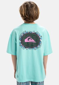 Camiseta oversized de color turquesa con un gráfico circular que presenta un logo de ola, un contorno de globo en negro y texto colorido en la parte posterior. Mangas cortas.