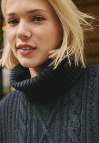 Dunkelblauer gestrickter Pullover mit hohem, geripptem Rollkragen und strukturierten Zopfmuster. Glatte Textur mit gemütlichem Erscheinungsbild.