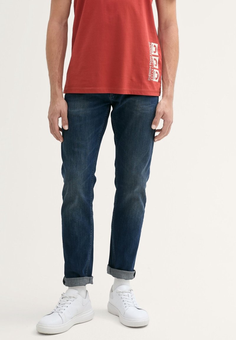 Červený bavlněný tílko s bílou grafickou potiskem, spárované s tmavě modrými džíny a bílými teniskami s texturovaným povrchem.