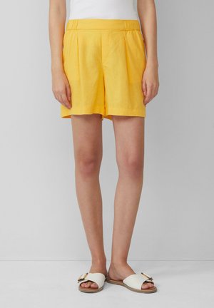 Shorts en lin jaune avec un design plissé et une taille élastique. Associés à des sandales blanches plates avec un détail de nœud décoratif.
