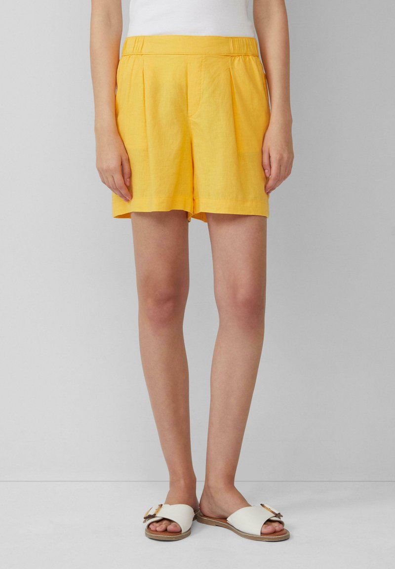 Yellow linen shorts with a pleated design and an elastic waistband. Paired with flat white sandals featuring a decorative bow detail.