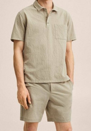 Mężczyzna w jasnobeżowej koszulce polo z krótkim rękawem z kieszenią na piersi oraz pasujących spodenkach do kolan, ręce luźno opuszczone, neutralne tło.