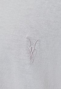 Světlě šedá látka s hladkou texturou, s malým vyšitým motivem kozí lebky v mírně tmavším odstínu šedé.