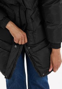 Svart puffjacka med dragkedja fram, tryckknappar och ribbade ärmar. Texturen är slät, med vadderade sektioner för isolering.