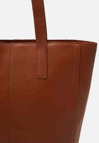 Bruine leren tote bag met een gestructureerde afwerking, platte handvatten en een gestructureerde vorm. Naaddetails zijn zichtbaar aan de zijkanten.