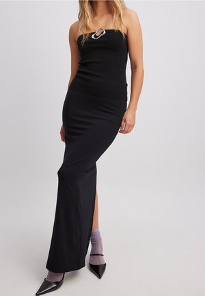 Woman in a strapless black dress with a silver brooch, wearing shiny black heels and purple socks, standing against a plain background.