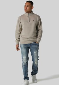 Beige zip-genser med høy krage, kombinert med slitte lys blå jeans og hvite joggesko, med en liten logo på brystet.