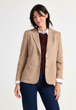Woman with dark hair wearing a beige blazer over a burgundy buttoned sweater and white shirt, paired with dark blue jeans, standing against white background.