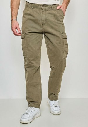 Homme portant un pantalon cargo vert olive et des baskets blanches, avec une main dans la poche, sur un fond clair uni.