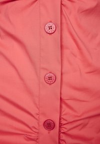 Korallivärinen kangas, jossa on sileä tekstuuri ja kolme näkyvää nappia: kaksi yksiväristä ja yksi kaiverretulla logolla, sijoitettuna pystysauman keskelle.