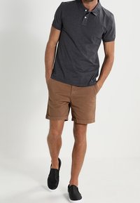 Polo shirt gris foncé avec deux boutons, associé à un short marron. La tenue est complétée par des chaussures noires à enfiler.