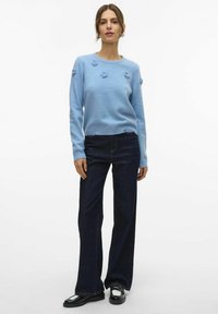 Vrouw in een lichtblauwe trui met bloemapplicaties, donkere wijde jeans en zwarte loafers, staand tegen een effen witte achtergrond.