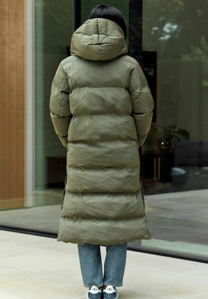 Manteau en duvet long et duveteux de couleur olive, avec une capuche, présentant une texture côtelée. Porté sur un jean en denim clair et des baskets.