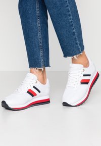 Baskets blanches avec des rayures rouges, noires et blanches, portées avec un jean bleu aux bords effilochés sur un sol et un fond clairs.