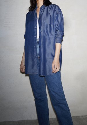 Personne portant une chemise bleue ample boutonnée sur un haut blanc, associée à un jean bleu, debout contre un mur clair uni.