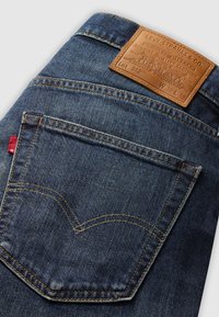 Blue denim jeans with a brown leather back patch. Features orange stitching and a distinctive v-shaped design on the back pocket.