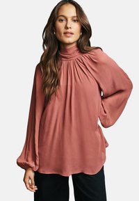 Blusa color rosa con maniche lunghe e voluminose, colletto alto e dettagli arricciati sulle spalle, realizzata in un tessuto leggero.