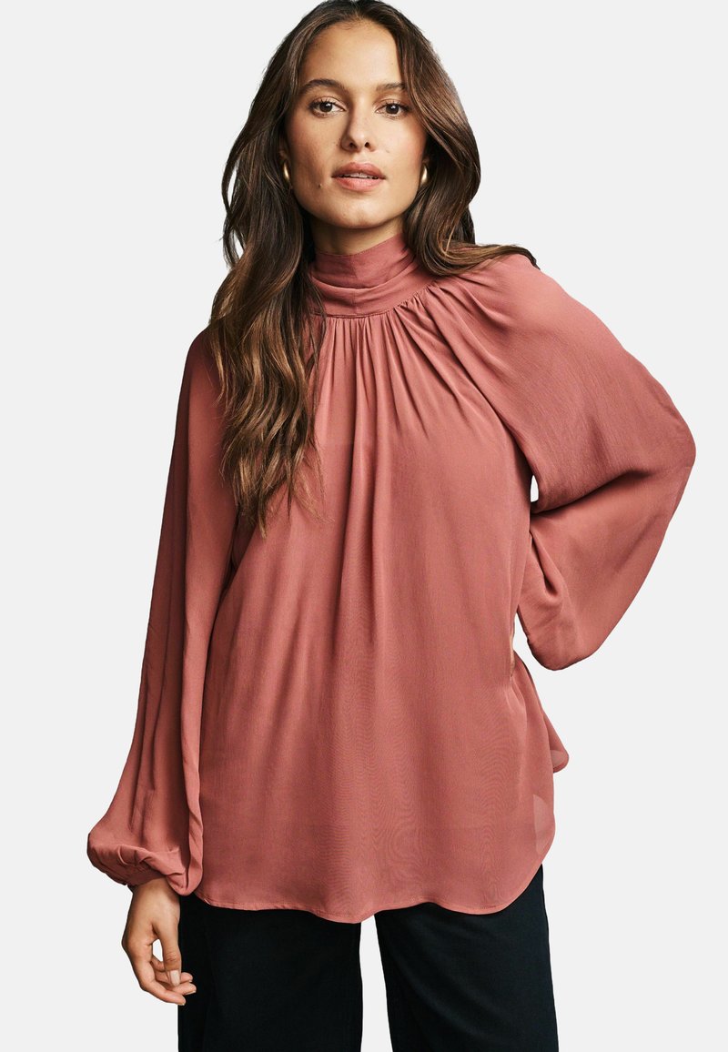 Blusa color rosa con maniche lunghe e voluminose, colletto alto e dettagli arricciati sulle spalle, realizzata in un tessuto leggero.