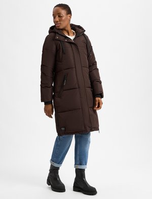 Brown padded long coat with a hood, featuring zip pockets and a textured finish. Paired with rolled-up jeans and ankle boots.