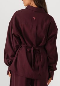 Burgundy oversized shirt with a tie waist feature, long sleeves, button cuffs, and a small embroidered logo on the upper back.