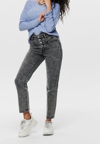 Hoge taille, lichtgrijze denimjeans met knoopsluiting, taps toelopende pijpen en vervaagde textuur, gecombineerd met een blauwe gebreide trui en witte sneakers.