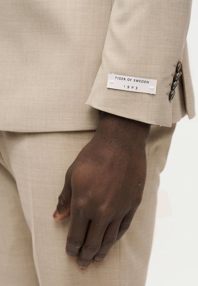 Κοντινό πλάνο σκουρόχρωμου χεριού δίπλα σε μανίκι μπεζ κοστουμιού με ετικέτα "Tiger of Sweden 1903" και τρία σκούρα κουμπιά στη μανσέτα.