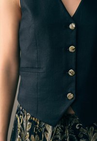 Dunkelgraue, maßgeschneiderte Weste mit V-Ausschnitt, fünf Messingknöpfen und Seitentaschen, kombiniert mit einem gemusterten Rock in Gold- und Marineblau-Tönen.