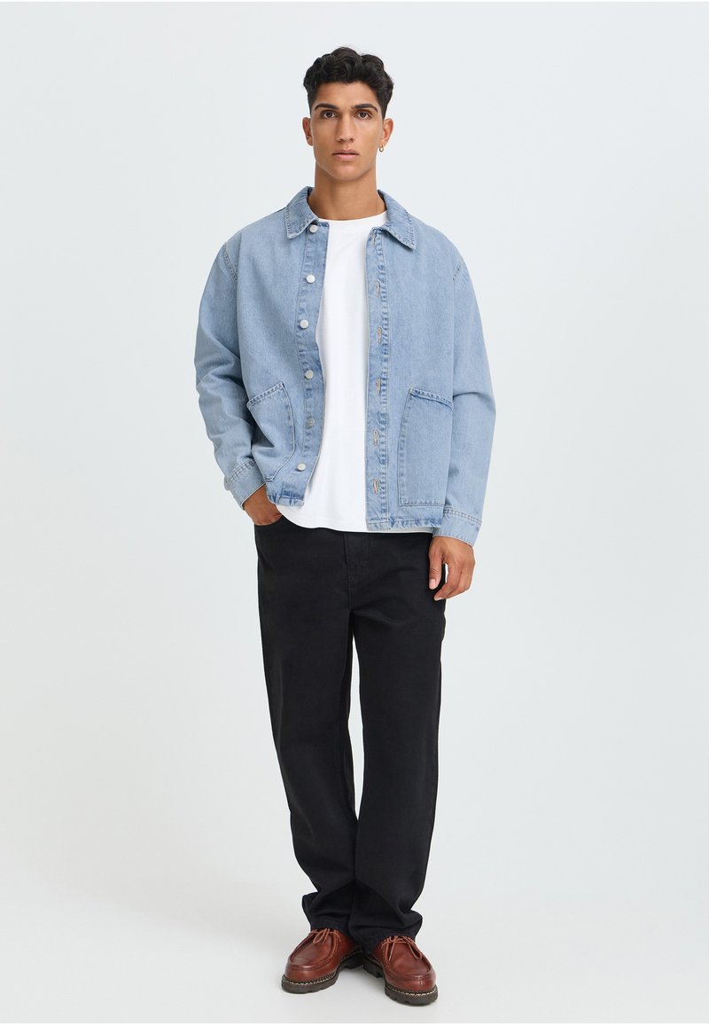 Jeune homme debout portant une veste en jean bleu clair, une chemise blanche, un pantalon noir et des chaussures marron sur un fond clair uni.