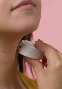 Hand-held white device being placed against the neck. Smooth surface, circular shape, minimalistic design, set against a pink background.