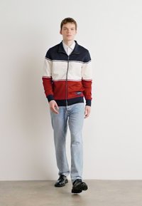 Giacca a zip in blu navy, crema e rosso con design a blocchi di colore. Taglio slim, polsini e colletto a costine. Jeans azzurri e scarpe nere.