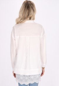 Witte blouse met lange mouwen, gemaakt van doorschijnende stof en een kantafwerking onderaan. De achterkant toont een plooi en een eenvoudig kraagontwerp.