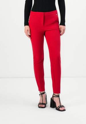 Vrouw in slim-fit rode broek, zwarte ribbed lange mouwen top en zwarte hoge hakken met enkelschoenen, staande op een witte vloer.