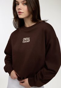 Młoda kobieta w ciemnobrązowej bluzie z napisem "KOCHAM KAWOWY KLUB" na środku, trzymająca rękami dół, na jednolitym jasnym tle.