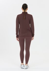 Maroon urheilu paita korkealla kauluksella ja pitkillä hihoilla, yhdistettynä vastaaviin marooniin leggingseihin. Tasainen materiaali ja hyvin istuva muotoilu, jota täydentävät harmaat lenkkarit.