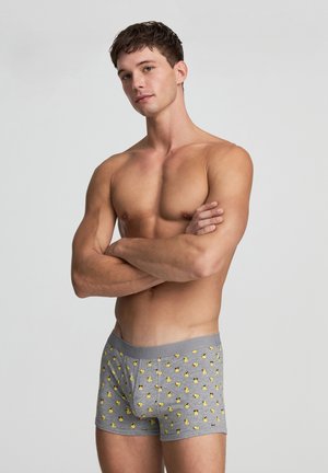 Uomo a torso nudo in piedi con le braccia incrociate, che indossa boxer grigi decorati con piccoli anatre di gomma gialla su uno sfondo semplice.
