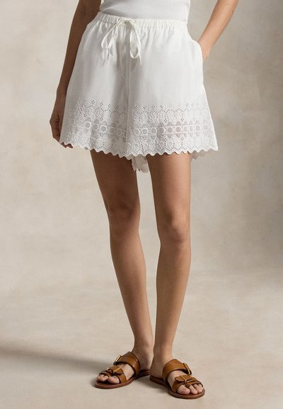 Γυναίκα που φοράει λευκά σορτς από δαντέλα eyelet με κορδόνι στη μέση και καφέ σανδάλια με αγκράφα, όρθια με το ένα χέρι στην τσέπη.