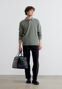 Άνδρας με γκρι μακρυμάνικη πόλο μπλούζα, μαύρο παντελόνι και παπούτσια περπατά κρατώντας μια μαύρη δερμάτινη τσάντα duffel στο δεξί του χέρι.