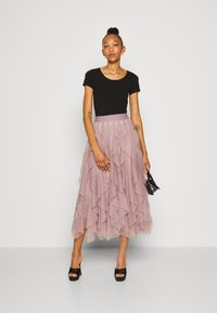 Haut noir ajusté associé à une jupe maxi en tulle rose à couches et volants. Le modèle porte des talons plats noirs et tient un petit sac à main noir.
