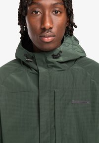Green hooded jacket with textured surface, featuring a front zipper, adjustable drawstrings, and a logo patch on the chest.