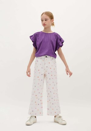 Chemise violette à manches courtes avec des manches volantes, associée à un pantalon large blanc avec un motif de fleurs multicolores. Baskets blanches.