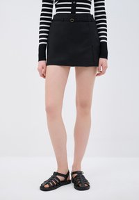 Gonna mini in pelle nera con uno spacco laterale, completata da una cintura sottile con fibbia circolare. Abbinata a sandali neri con fasce.