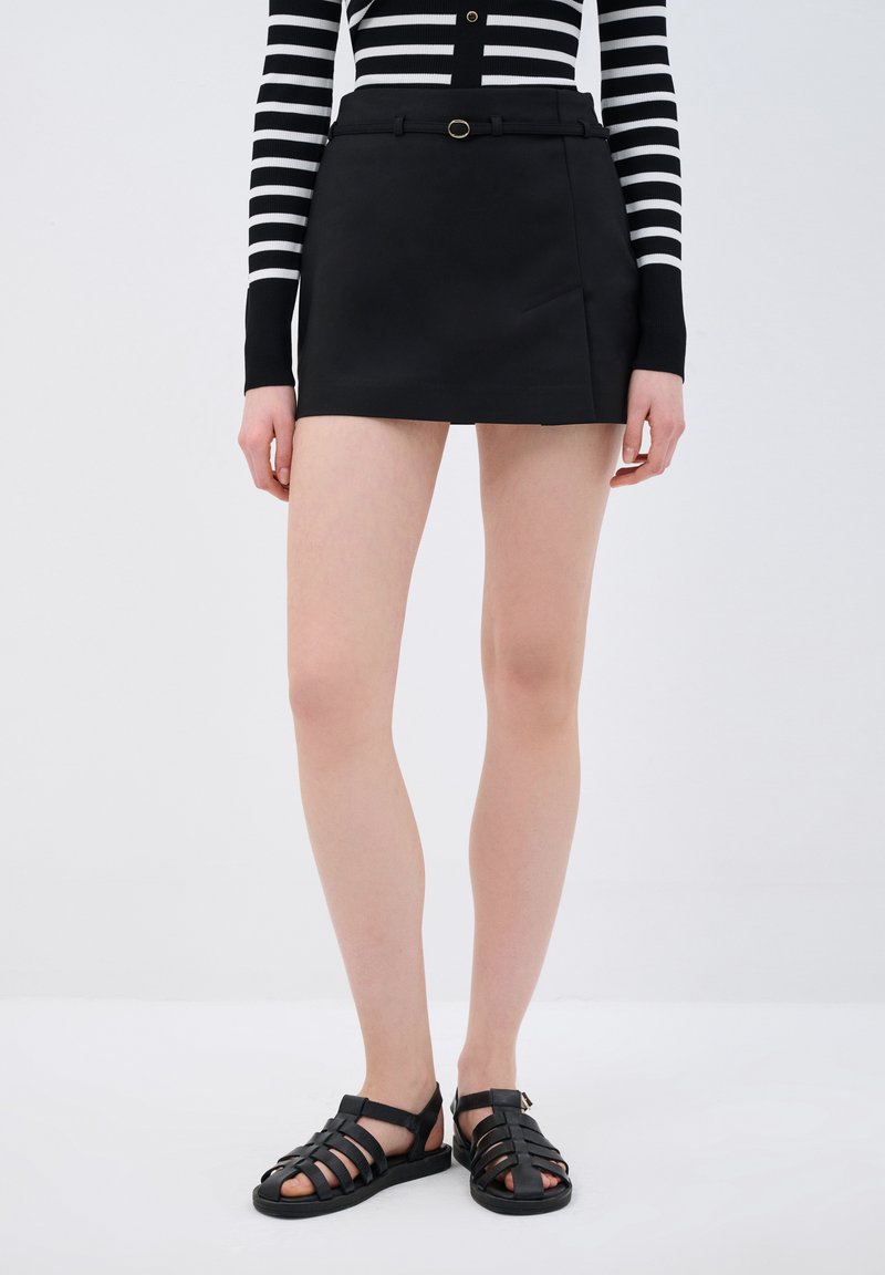 Gonna mini in pelle nera con uno spacco laterale, completata da una cintura sottile con fibbia circolare. Abbinata a sandali neri con fasce.