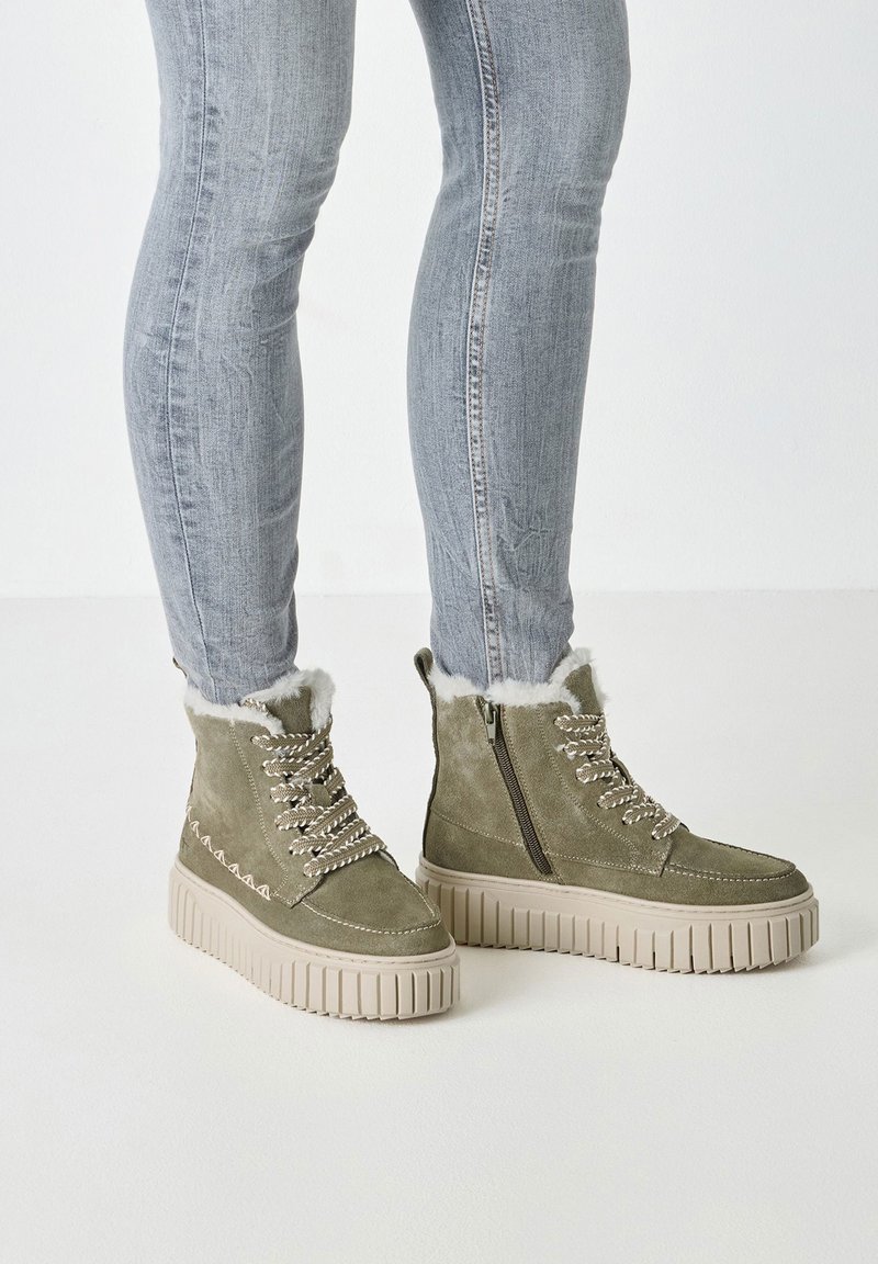 Persoon gekleed in lichtgrijze skinny jeans en olijfgroene suède winterlaarzen met witte bontvoering en dikke beige zolen.