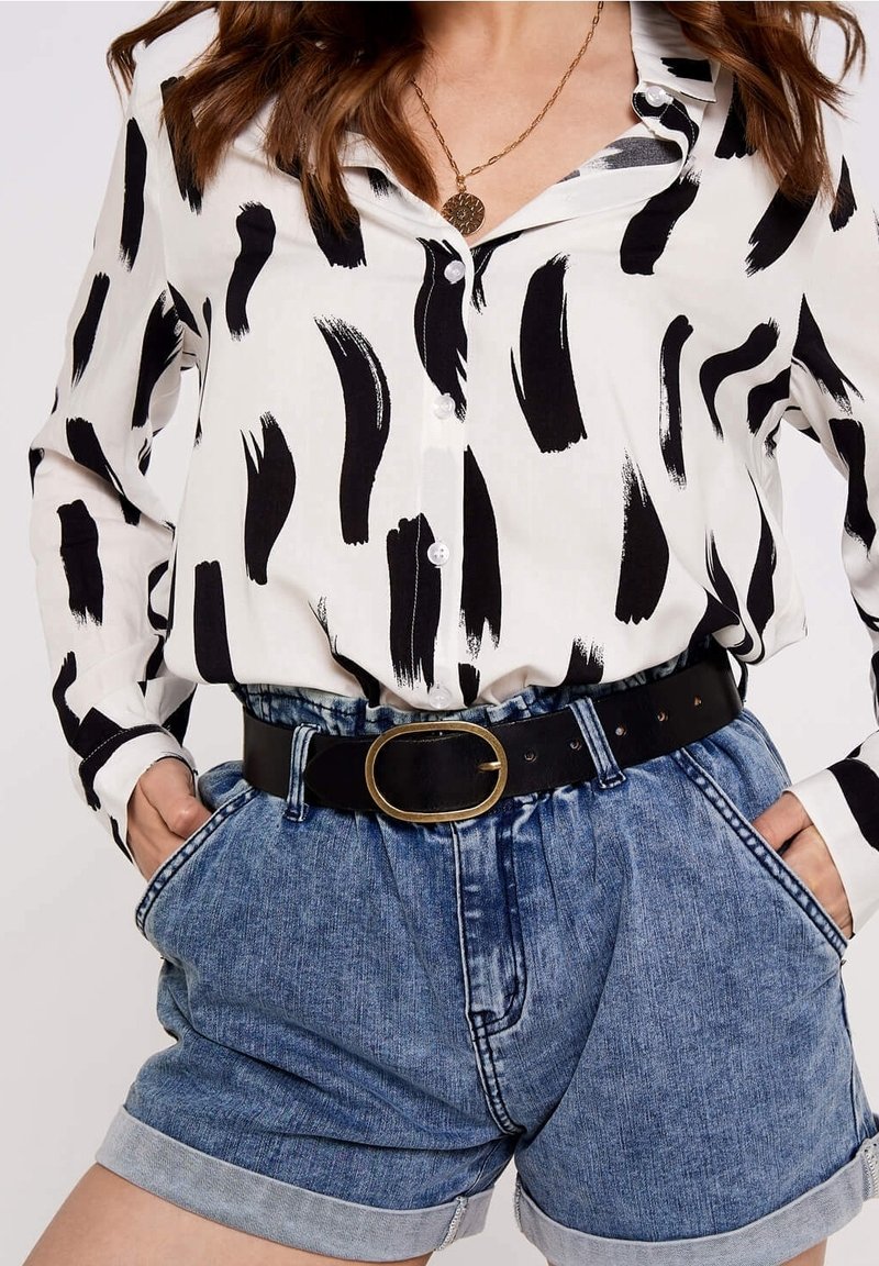 Blouse blanche avec un motif de coups de pinceau noirs, boutonnée sur le devant. Associée à un short en denim bleu clair taille haute et une ceinture noire avec boucle dorée.