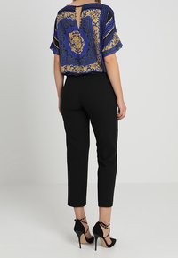 Frau in schwarzen, schmalen Hosen, schwarzen, hochhackigen Schuhen mit Knöchelriemen und einem kurzärmeligen blauen Oberteil mit goldenen barocken Mustern, steht mit dem Rücken zur Kamera.