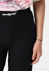 Black ribbed leggings with a high waistband featuring white "lowlights" branding. Close-up showing hand with a tattoo against a neutral background.