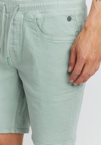 Pantaloni scurți din bumbac de culoare verde deschis, cu talie elastică și șnur. Au buzunare laterale și o mică insignă cu logo pe partea din față.