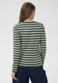 Sieviete ar gariem brūniem matiem, tērpta zaļi un baltās svītrainās garām piedurknēm kretē un tumši zilās biksēs, skatīta no aizmugures.