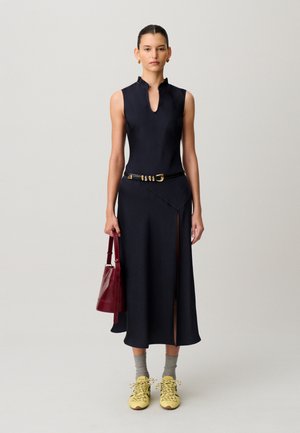 Mujer con vestido negro sin mangas con abertura lateral, cinturón negro, calcetines grises, zapatillas amarillas, sosteniendo un bolso rojo sobre un fondo liso.