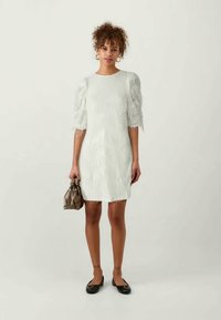 Balta suknelė su pūkuotu tekstūro, trumpomis rankovėmis ir tiesiu kirpimu. Modelis laiko mažą rudąRankinę ir dėvi juodus butus. Fonas neutralus.